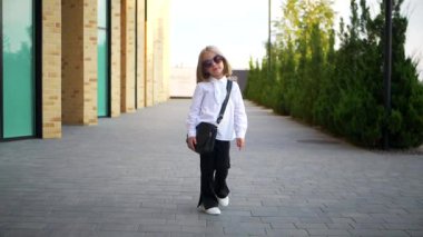 Beyaz gömlekli ve siyah pantolonlu genç bir kız kaldırımda duruyor, gülümsüyor ve kameraya bakıyor. Şehir kaldırımında kendine güvenen bir duruşu olan beyaz bluz ve siyah pantolon giyen küçük bir kız.