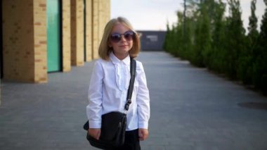 Beyaz gömlekli ve siyah pantolonlu genç bir kız kaldırımda duruyor, gülümsüyor ve kameraya bakıyor. Şehir kaldırımında kendine güvenen bir duruşu olan beyaz bluz ve siyah pantolon giyen küçük bir kız.