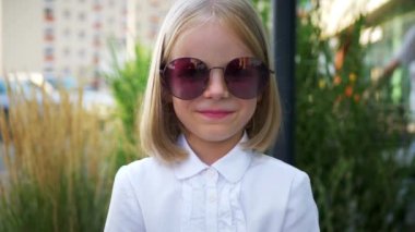 Beyaz gömlekli ve siyah pantolonlu genç bir kız kaldırımda duruyor, gülümsüyor ve kameraya bakıyor. Şehir kaldırımında kendine güvenen bir duruşu olan beyaz bluz ve siyah pantolon giyen küçük bir kız.