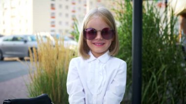 Beyaz gömlekli ve siyah pantolonlu genç bir kız kaldırımda duruyor, gülümsüyor ve kameraya bakıyor. Şehir kaldırımında kendine güvenen bir duruşu olan beyaz bluz ve siyah pantolon giyen küçük bir kız.