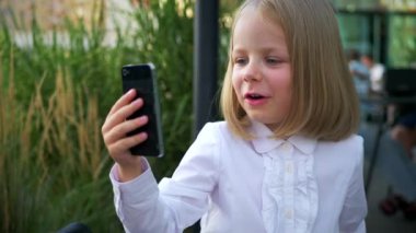Güneş gözlüğü takan, cep telefonuyla konuşan, iş merkezinin yakınında kendinden emin ve neşeli görünen küçük bir kız. Video iletişimi, Skype. Telefonuyla selfie çeken bir kız.