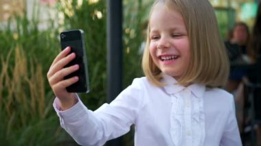 Güneş gözlüğü takan, cep telefonuyla konuşan, iş merkezinin yakınında kendinden emin ve neşeli görünen küçük bir kız. Video iletişimi, Skype. Telefonuyla selfie çeken bir kız.