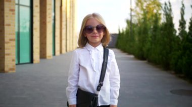 Beyaz gömlekli ve siyah pantolonlu genç bir kız kaldırımda duruyor, gülümsüyor ve kameraya bakıyor. Şehir kaldırımında kendine güvenen bir duruşu olan beyaz bluz ve siyah pantolon giyen küçük bir kız.