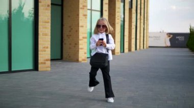 Güneş gözlüklü küçük mutlu bir kız cep telefonunu tutarken gülümsüyor. Genç bir kız şık güneş gözlüğü takıyor, akıllı telefonuyla oynuyor ve çok mutlu. Güneş gözlüklü neşeli bir kız, telefonuyla anları yakalıyor..