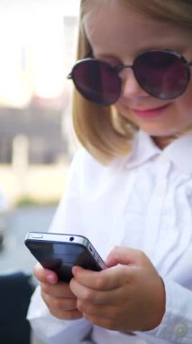 Dikey, şık güneş gözlüğü takan genç bir kız akıllı telefonuyla oynuyor ve çok mutlu. Güneş gözlüklü mutlu bir kız telefonunda mesaj atıyor. İş kadını, internette iletişim.