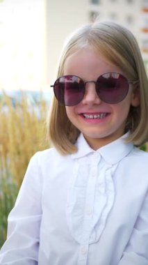 Dikey, beyaz gömlekli ve siyah pantolonlu genç bir kız kaldırımda duruyor, gülümsüyor ve kameraya bakıyor. Şehir kaldırımında kendine güvenen bir duruşu olan beyaz bluz ve siyah pantolon giyen küçük bir kız.