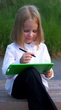 Dikey, bir bankta oturan, tabletine odaklanmış genç bir kız. Bir kız parkta bankta otururken mutlu ve tabletinin tadını çıkarıyor. İş kadını. Tablet cihazıyla meşgul bir çocuk bankta oturuyor..