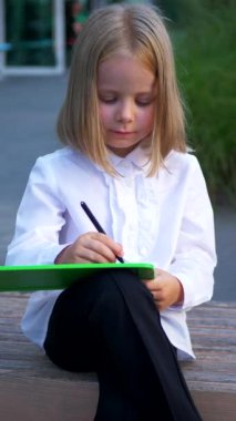 Dikey, bir bankta oturan, tabletine odaklanmış genç bir kız. Bir kız parkta bankta otururken mutlu ve tabletinin tadını çıkarıyor. İş kadını. Tablet cihazıyla meşgul bir çocuk bankta oturuyor..