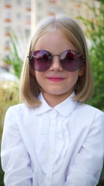 Dikey, beyaz gömlekli ve siyah pantolonlu genç bir kız kaldırımda duruyor, gülümsüyor ve kameraya bakıyor. Şehir kaldırımında kendine güvenen küçük bir kız, beyaz bir bluz giymiş. 