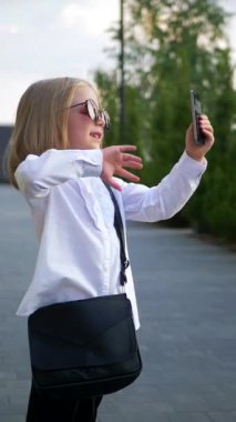 Dikey. Güneş gözlüğü takan, cep telefonuyla konuşan, iş merkezinin yakınında kendinden emin ve neşeli görünen küçük bir kız. Video iletişimi, Skype. Mutlu bir kız telefonuyla selfie çekiyor.
