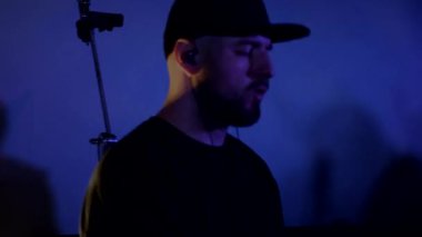 Davulcu cool rock konserini çalar. Bir adam sahnede enerjik bir şekilde davul çalıyor arkasında parlak ışıklar yanıyor. Canlı performans sergileyen bir davulcu. Etrafı renkli sahne ışıklarıyla çevrili. Parlak ışıklarla davul çalan bir adam 