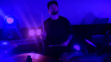 Davulcu cool rock konserini çalar. Bir adam sahnede enerjik bir şekilde davul çalıyor arkasında parlak ışıklar yanıyor. Canlı performans sergileyen bir davulcu, renkli sahne ışıklarıyla çevrili..