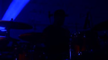 Davulcu cool rock konserini çalar. Bir adam sahnede enerjik bir şekilde davul çalıyor arkasında parlak ışıklar yanıyor. Canlı performans sergileyen bir davulcu, renkli sahne ışıklarıyla çevrili..