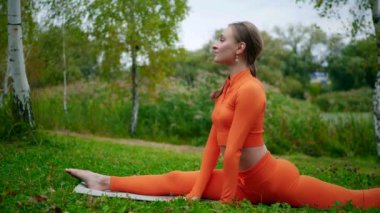Yoga yaparken parkta gerinen turuncu kıyafetli bir kadın. Esneklik eğitimi, Limbering rutinleri, hareket alıştırmaları, müşterek seferberlik tatbikatları, esneklik egzersizleri, fiziksel egzersizler, spor egzersizleri yapan bir kız.