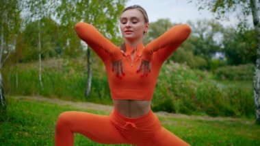 Yoga yaparken parkta gerinen turuncu kıyafetli bir kadın. Esneklik eğitimi, Limbering rutinleri, hareket alıştırmaları, müşterek seferberlik tatbikatları, esneklik egzersizleri, fiziksel egzersizler, spor egzersizleri yapan bir kız.