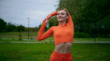 Yoga yaparken parkta gerinen turuncu kıyafetli bir kadın. Esneklik eğitimi, Limbering rutinleri, hareket alıştırmaları, müşterek seferberlik tatbikatları, esneklik egzersizleri, fiziksel egzersizler, spor egzersizleri yapan bir kız.