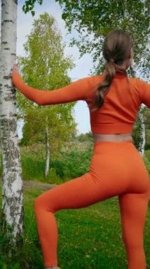 Dikey. Yoga yaparken parkta gerinen turuncu kıyafetli bir kadın. Bir kadın esneme egzersizleri yapar. Esneklik eğitimi, Limbering rutinleri, Hareket çeşitliliği tatbikatları, Ortak seferberlik tatbikatları, esneklik egzersizleri,