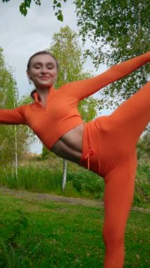 Dikey. Yoga yaparken parkta gerinen turuncu kıyafetli bir kadın. Bir kadın esneme egzersizleri yapar. Esneklik eğitimi, Limbering rutinleri, Hareket çeşitliliği tatbikatları, Ortak seferberlik tatbikatları, esneklik egzersizleri,