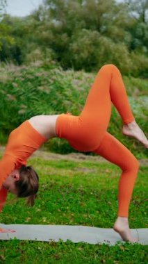 Dikey. Yoga yaparken parkta gerinen turuncu kıyafetli bir kadın. Bir kadın esneme egzersizleri yapar. Esneklik eğitimi, Limbering rutinleri, Hareket çeşitliliği tatbikatları, Ortak seferberlik tatbikatları, esneklik egzersizleri,