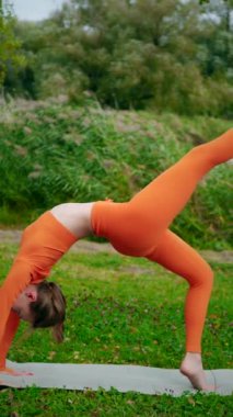 Dikey. Yoga yaparken parkta gerinen turuncu kıyafetli bir kadın. Bir kadın esneme egzersizleri yapar. Esneklik eğitimi, Limbering rutinleri, Hareket çeşitliliği tatbikatları, Ortak seferberlik tatbikatları, esneklik egzersizleri,