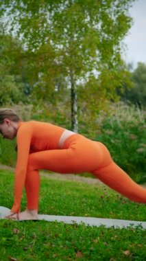 Dikey. Yoga yaparken parkta gerinen turuncu kıyafetli bir kadın. Bir kadın esneme egzersizleri yapar. Esneklik eğitimi, Limbering rutinleri, Hareket çeşitliliği tatbikatları, Ortak seferberlik tatbikatları, esneklik egzersizleri,