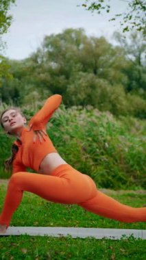 Dikey. Yoga yaparken parkta gerinen turuncu kıyafetli bir kadın. Bir kadın esneme egzersizleri yapar. Esneklik eğitimi, Limbering rutinleri, Hareket çeşitliliği tatbikatları, Ortak seferberlik tatbikatları, esneklik egzersizleri,