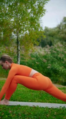 Dikey. Yoga yaparken parkta gerinen turuncu kıyafetli bir kadın. Bir kadın esneme egzersizleri yapar. Esneklik eğitimi, Limbering rutinleri, Hareket çeşitliliği tatbikatları, Ortak seferberlik tatbikatları, esneklik egzersizleri,