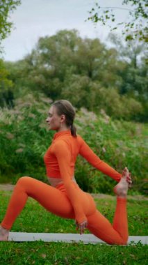 Dikey. Yoga yaparken parkta gerinen turuncu kıyafetli bir kadın. Bir kadın esneme egzersizleri yapar. Esneklik eğitimi, Limbering rutinleri, Hareket çeşitliliği tatbikatları, Ortak seferberlik tatbikatları, esneklik egzersizleri,