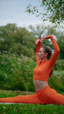 Dikey. Yoga yaparken parkta gerinen turuncu kıyafetli bir kadın. Bir kadın esneme egzersizleri yapar. Esneklik eğitimi, Limbering rutinleri, Hareket çeşitliliği tatbikatları, Ortak seferberlik tatbikatları, esneklik egzersizleri,