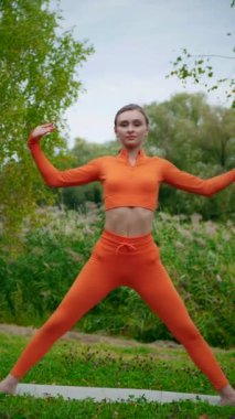 Dikey. Yoga yaparken parkta gerinen turuncu kıyafetli bir kadın. Bir kadın esneme egzersizleri yapar. Esneklik eğitimi, Limbering rutinleri, Hareket çeşitliliği tatbikatları, Ortak seferberlik tatbikatları, esneklik egzersizleri,