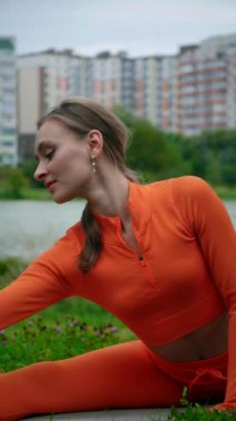 Dikey. Yoga yaparken parkta gerinen turuncu kıyafetli bir kadın. Bir kadın esneme egzersizleri yapar. Esneklik eğitimi, Limbering rutinleri, Hareket çeşitliliği tatbikatları, Ortak seferberlik tatbikatları, esneklik egzersizleri,