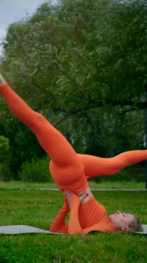 Dikey. Yoga yaparken parkta gerinen turuncu kıyafetli bir kadın. Bir kadın esneme egzersizleri yapar. Esneklik eğitimi, Limbering rutinleri, Hareket çeşitliliği tatbikatları, Ortak seferberlik tatbikatları, esneklik egzersizleri,