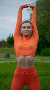 Dikey. Yoga yaparken parkta gerinen turuncu kıyafetli bir kadın. Bir kadın esneme egzersizleri yapar. Esneklik eğitimi, Limbering rutinleri, Hareket çeşitliliği tatbikatları, Ortak seferberlik tatbikatları, esneklik egzersizleri,