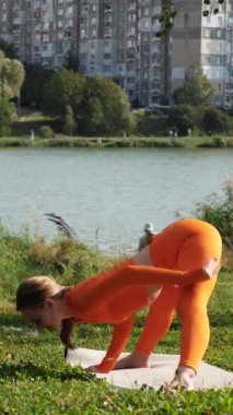 Dikey. Yoga yaparken parkta gerinen turuncu kıyafetli bir kadın. Bir kadın esneme egzersizleri yapar. Esneklik eğitimi, Limbering rutinleri, Hareket çeşitliliği tatbikatları, Ortak seferberlik tatbikatları, esneklik egzersizleri,