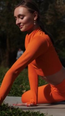 Dikey. Yoga yaparken parkta gerinen turuncu kıyafetli bir kadın. Bir kadın esneme egzersizleri yapar. Esneklik eğitimi, Limbering rutinleri, Hareket çeşitliliği tatbikatları, Ortak seferberlik tatbikatları, esneklik egzersizleri,