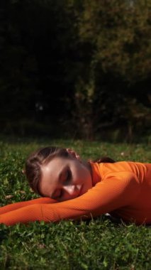 Dikey. Yoga yaparken parkta gerinen turuncu kıyafetli bir kadın. Bir kadın esneme egzersizleri yapar. Esneklik eğitimi, Limbering rutinleri, Hareket çeşitliliği tatbikatları, Ortak seferberlik tatbikatları, esneklik egzersizleri,