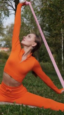 Dikey. Yoga yaparken parkta gerinen turuncu kıyafetli bir kadın. Bir kadın esneme egzersizleri yapar. Esneklik eğitimi, Limbering rutinleri, Hareket çeşitliliği tatbikatları, Ortak seferberlik tatbikatları, esneklik egzersizleri,