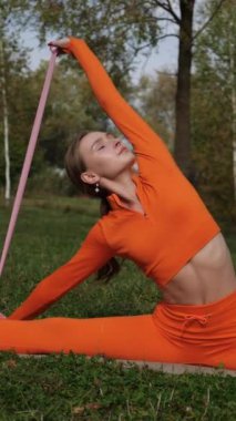 Dikey. Yoga yaparken parkta gerinen turuncu kıyafetli bir kadın. Bir kadın esneme egzersizleri yapar. Esneklik eğitimi, Limbering rutinleri, Hareket çeşitliliği tatbikatları, Ortak seferberlik tatbikatları, esneklik egzersizleri,