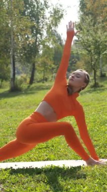 Dikey. Yoga yaparken parkta gerinen turuncu kıyafetli bir kadın. Bir kadın esneme egzersizleri yapar. Esneklik eğitimi, Limbering rutinleri, Hareket çeşitliliği tatbikatları, Ortak seferberlik tatbikatları, esneklik egzersizleri,