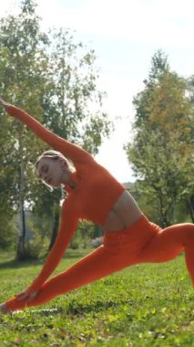 Dikey. Yoga yaparken parkta gerinen turuncu kıyafetli bir kadın. Bir kadın esneme egzersizleri yapar. Esneklik eğitimi, Limbering rutinleri, Hareket çeşitliliği tatbikatları, Ortak seferberlik tatbikatları, esneklik egzersizleri,