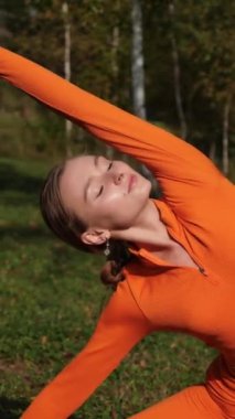 Dikey. Yoga yaparken parkta gerinen turuncu kıyafetli bir kadın. Bir kadın esneme egzersizleri yapar. Esneklik eğitimi, Limbering rutinleri, Hareket çeşitliliği tatbikatları, Ortak seferberlik tatbikatları, esneklik egzersizleri,