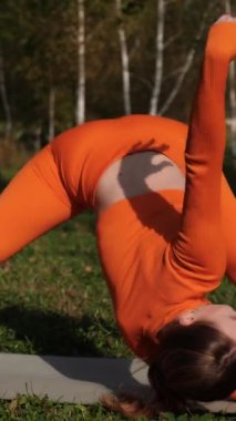 Dikey. Yoga yaparken parkta gerinen turuncu kıyafetli bir kadın. Bir kadın esneme egzersizleri yapar. Esneklik eğitimi, Limbering rutinleri, Hareket çeşitliliği tatbikatları, Ortak seferberlik tatbikatları, esneklik egzersizleri,