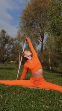 Dikey. Yoga yaparken parkta gerinen turuncu kıyafetli bir kadın. Bir kadın esneme egzersizleri yapar. Esneklik eğitimi, Limbering rutinleri, Hareket çeşitliliği tatbikatları, Ortak seferberlik tatbikatları, esneklik egzersizleri,