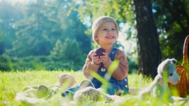 Sonbahar parkı sahnesi; yakut kırmızısı bir elma tutan bir çocuk, doğanın güzelliğiyle çevrili bir battaniyenin üzerinde oturur. Küçük bir tanesi gülümsüyor, canlı yeşil çimenlerin arasına yerleşmiş, tatlı bir ziyafetin tadını çıkarıyor, altın güneş ışığıyla yıkanmış. Parkta piknik zamanı; bir çocuk,