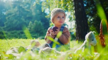 Sonbahar parkı sahnesi; yakut kırmızısı bir elma tutan bir çocuk, doğanın güzelliğiyle çevrili bir battaniyenin üzerinde oturur. Küçük bir tanesi gülümsüyor, canlı yeşil çimenlerin arasına yerleşmiş, tatlı bir ziyafetin tadını çıkarıyor, altın güneş ışığıyla yıkanmış. Parkta piknik zamanı; bir çocuk,