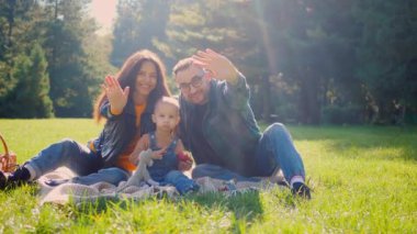 Kahkaha ve sevgi dolu güneşli bir öğleden sonra pikniği. Aile dışarıda eğleniyor ve kameraya el sallıyor. Sıcak güneş ışığı, mutlu yüzler ve dost dalgalar. Mükemmel bir aile anı. Doğanın kucakladığı bir aile. Neşeli bağlantı 