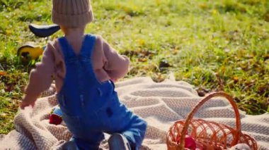 Sonbahar parkı sahnesi; yakut kırmızısı bir elma tutan bir çocuk, doğanın güzelliğiyle çevrili bir battaniyenin üzerinde oturur. Küçük bir tanesi gülümsüyor, canlı yeşil çimenlerin arasına yerleşmiş, tatlı bir ziyafetin tadını çıkarıyor, altın güneş ışığıyla yıkanmış. Parkta piknik zamanı; bir çocuk,