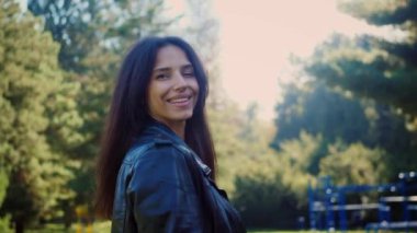 Sıcak bir gülüşü olan, bir parkın altın ışığında yıkanan bir kadın. Hışırdayan yaprakların ve canlı renklerin arasında bir mutluluk anı. Bir kadının açık hava portresi, sıcak ve cezbedici bir gülümseme. Uzun koyu renk saçlar, parlak turuncu bir kazak.