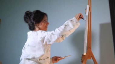 Gülümseyen bir sanatçı sehpaya resim çizer. Mutlu bir kadın tuvalde bir resim yaratıyor. Kadın, sanat stüdyosundaki beyaz kağıda gülüyor ve soyutlama çiziyor..