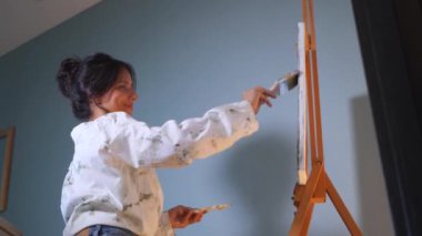 Gülümseyen bir sanatçı sehpaya resim çizer. Mutlu bir kadın tuvalde bir resim yaratıyor. Kadın, sanat stüdyosundaki beyaz kağıda gülüyor ve soyutlama çiziyor..